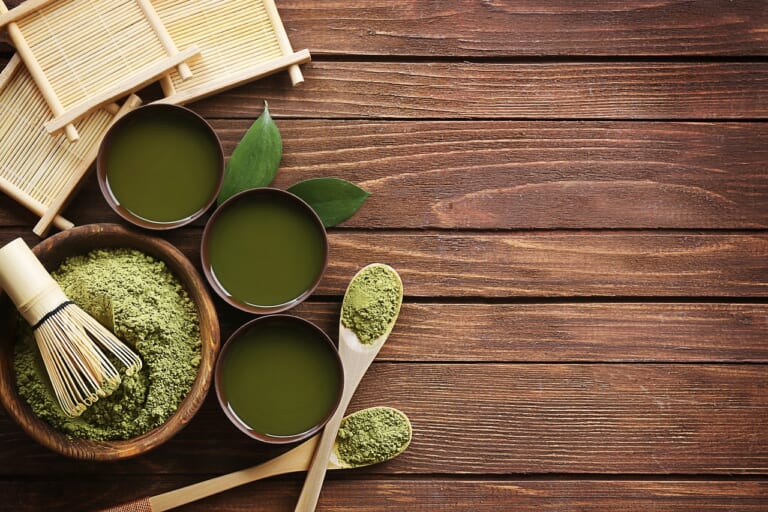 Matcha powder, a bamboo whisk and three bowls of green tea on a wooden table