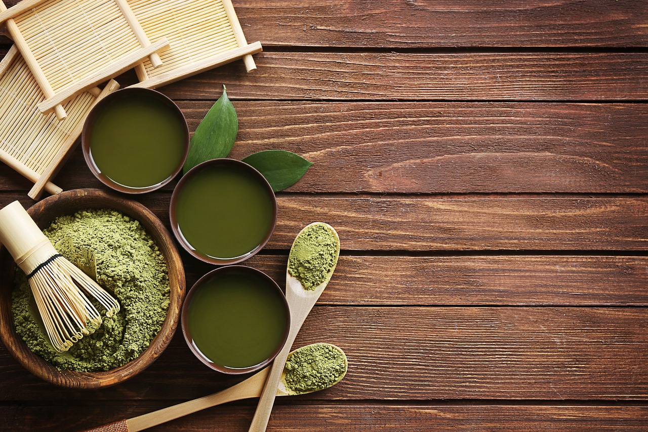 Matcha powder, a bamboo whisk and three bowls of green tea on a wooden table