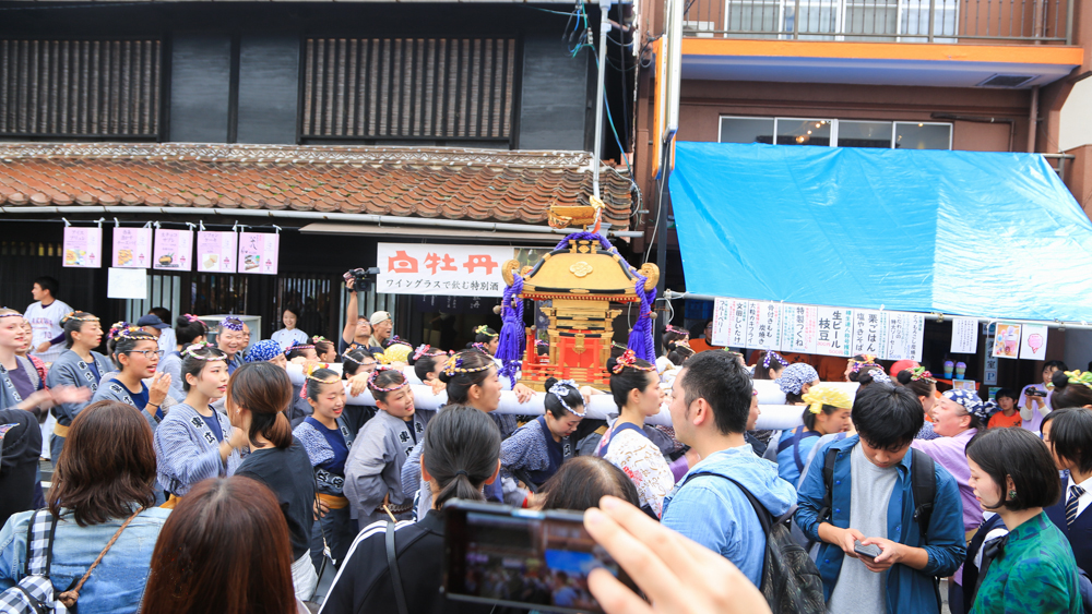 A portable shrine and crowds passing brewery buildings at Saijo Sake Festival