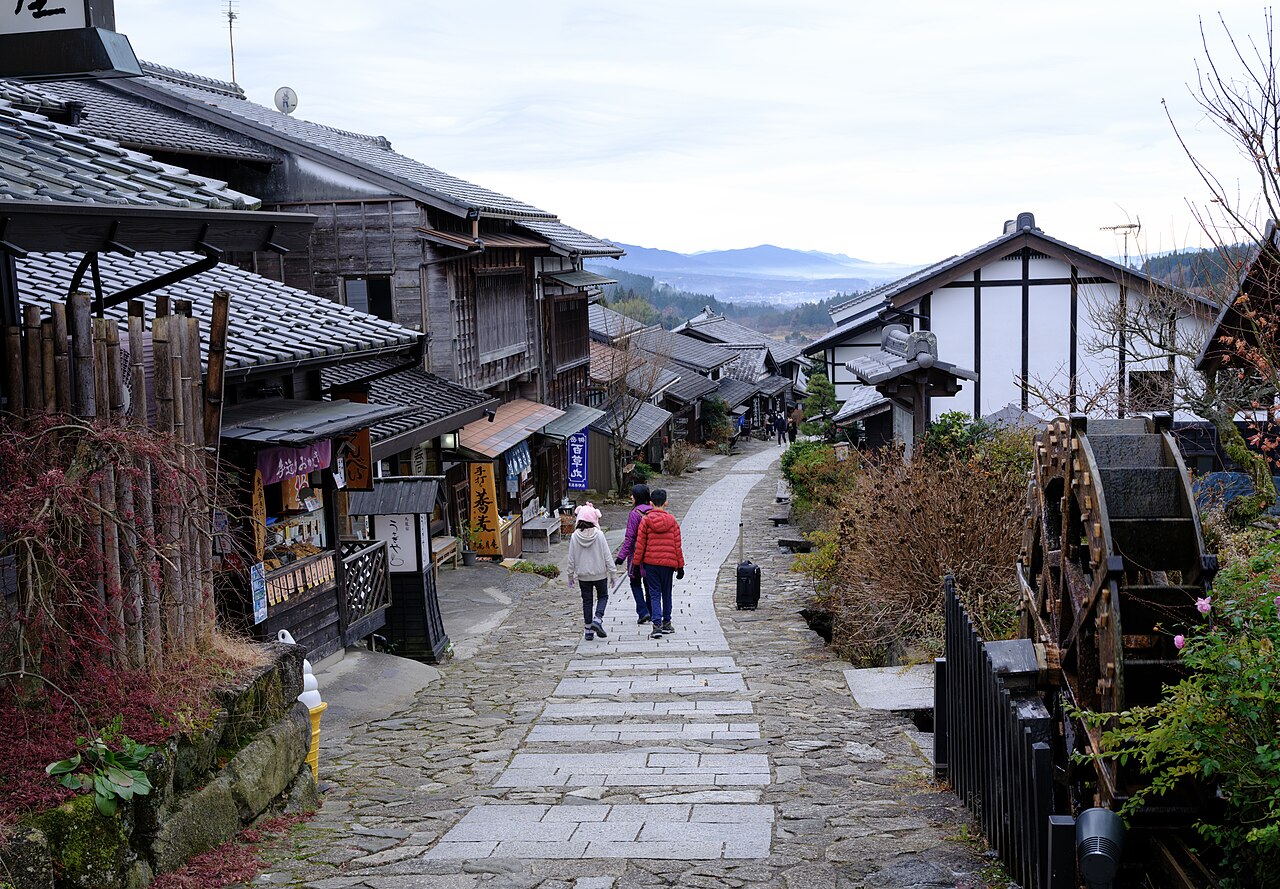 Walkers on Magome’s stone-paved slope beside wooden houses and a waterwheel